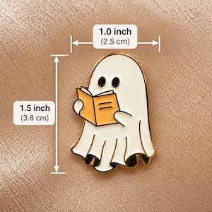 May include: A white ghost enamel pin with black eyes holding an open orange book. The pin features gold-toned metal edges and measures 1.0 inch in width by 1.5 inches in height, as indicated by the visible dimension labels on the image.