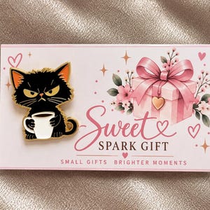 Puede incluir: Un pin de esmalte de gato negro con orejas interiores naranjas sosteniendo una taza de café blanca, sujeto a una tarjeta rectangular con una ilustración de caja de regalo rosa, flores y el texto Sweet SPARK GIFT SMALL GIFTS BRIGHTER MOMENTS.