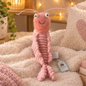 May include: A pink plush shrimp toy with a fuzzy head, corduroy-textured body, and two long antennae. The toy features two white eyes with black pupils, a stitched smile, and two attached paper tags, one of which displays the Jellycat brand logo.
