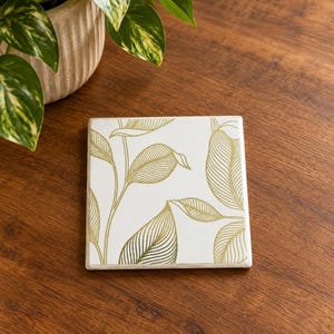 May include: A square ceramic coaster featuring a white surface with a gold-colored line art pattern of various leaves and stems. The coaster has a smooth finish and is positioned flat, displaying a detailed botanical illustration in metallic gold ink.