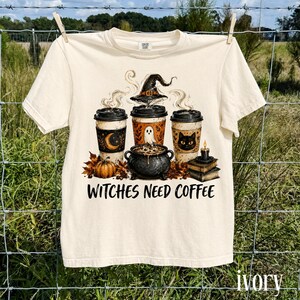 Peut inclure: Un t-shirt à manches courtes couleur ivoire avec un motif représentant trois tasses de café, un chaudron, une citrouille, une pile de livres et une bougie. Le dessin inclut un chapeau de sorcière, un fantôme et un chat. Texte WITCHES NEED COFFEE.