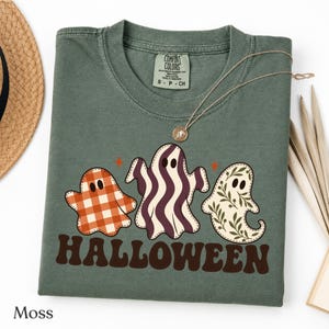 Könnte beinhalten: Ein moosgrünes T-Shirt mit Rundhalsausschnitt und einer Grafik von drei Geistern über dem Wort HALLOWEEN in braunen Buchstaben. Der linke Geist hat ein orange-weißes Karomuster, der mittlere violette Wellenstreifen und der rechte einen grünen Blätterdruck.