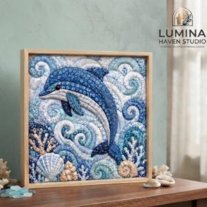 Puede incluir: Una pieza de arte textil cuadrada enmarcada que muestra un delfín azul nadando entre olas arremolinadas. El diseño está compuesto por telas con motivos florales en tonos azules y blancos, con detalles de coral y conchas texturizadas.
