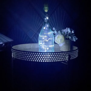 May include: A clear, ribbed glass bottle illuminated from within by a string of small, bright white LED fairy lights. The bottle is placed on a round, metallic tray with a perforated side pattern, next to a small vase containing white flowers.