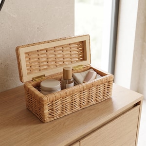 Puede incluir: Una caja de almacenamiento rectangular de ratán trenzado con tapa abatible y bisagras de metal dorado. La caja abierta contiene un pequeño recipiente redondo con tapa beige, una botella de vidrio transparente con tapón de madera, una tela plegada y un peine de madera.