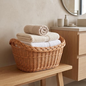 Pode incluir: Uma cesta de vime trançado castanho-claro contendo três toalhas dobradas e enroladas em tons de branco, creme e bege. A cesta apresenta uma borda trançada e está cheia de itens têxteis macios empilhados ordenadamente no recipiente.