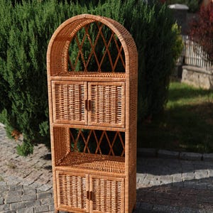 Puede incluir: Una unidad de almacenamiento alta de mimbre marrón claro con una parte superior arqueada y un patrón de celosía de diamantes. Incluye dos juegos de puertas dobles con pequeñas manijas de madera, dos estantes abiertos y textura tejida.