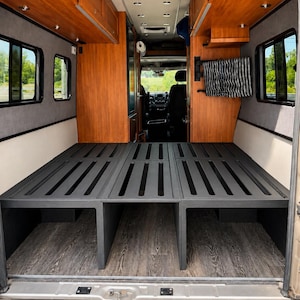 Può includere: L'interno di un camper con una struttura letto modulare grigio scuro a doghe. Il veicolo presenta pensili in legno, pannelli grigi alle pareti e un pavimento in legno scuro. Un tessuto a righe bianche e nere è appeso alla parete laterale.