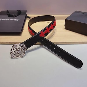 Può includere: Una cintura in pelle nera caratterizzata da una serie di applicazioni a forma di croce rossa lungo il cinturino e da una grande fibbia in metallo argentato lucido, ornata con dettagli a volute, presentata con scatola e borsa nere del marchio.