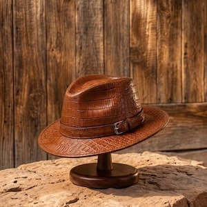 May include: A brown fedora hat with a textured crocodile skin pattern. The hat features a matching brown band around the crown, secured with a small metal buckle on the side. The brim is slightly upturned, and the crown has a central crease.