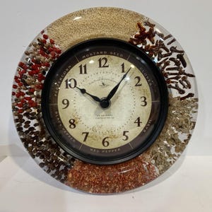 May include: A round wall clock encased in clear resin featuring embedded dried spices including peppercorns, mustard seeds, cloves, basil, and red pepper. The clock face has a cream background with black numerals and text labeling the spices.