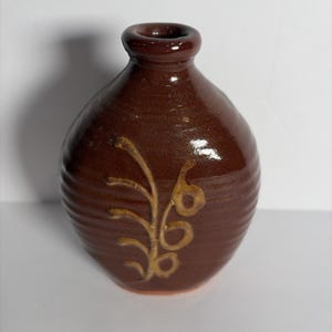 May include: A small, round, dark brown ceramic vase with a glossy finish and a narrow neck. The body of the vase features horizontal ridges and a light tan, hand-painted botanical design of a stem with leaves and berries on the front surface.