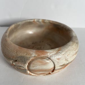 May include: A round ceramic bowl with a marbled finish in shades of beige, white, and terracotta. The exterior features a textured surface with a prominent circular indentation. The interior base displays the stamped text "MADE BY RON A".