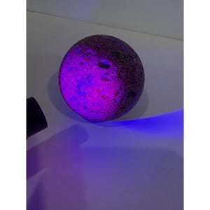 May include: A spherical rock with a porous, textured surface that glows with vibrant purple and blue light when illuminated by a handheld ultraviolet flashlight. The object displays a dark, pitted exterior that reacts to the light source.