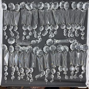 May include: A collection of clear, faceted glass chandelier crystals arranged in multiple strands. Each strand features a combination of octagonal beads and elongated rectangular prisms connected by small metal rings, creating a reflective design.