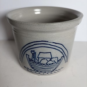 May include: A light gray ceramic crock featuring a dark blue embossed design of a boat carrying animals under a rainbow. The vessel has a wide, flared rim and a speckled texture throughout the exterior surface, with a smooth, rounded top edge.