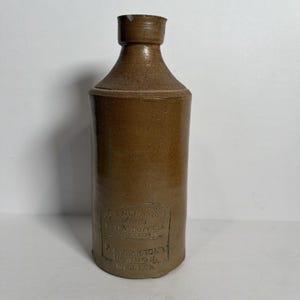 May include: A brown stoneware bottle with a cylindrical body and a narrow neck. The front features an embossed stamp that reads: J. BOURNE & SON, PATENTEES, DENBY POTTERY, NEAR DERBY, R.A.J. ARNOLD, LONDON, ENGLAND.