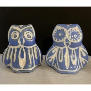 May include: Two ceramic owl figurines featuring a blue glaze with raised white outlines. The left owl has simple circular eyes, while the right owl has detailed gear-like patterns around its eyes. Both figures display stylized wing designs.