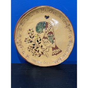 May include: A round, tan ceramic plate featuring a hand-painted illustration of a bird perched on a flowering branch. The text reads: A treasure; a Brother, a comfort; a Friend is both. - Ben Franklin. A small dark heart is painted above the bird.