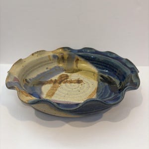 May include: A round ceramic pie dish with a ruffled, scalloped edge. The interior features a glaze combination of deep blue, cream, and tan, with a visible spiral texture and a cross-like mark in the center of the base of the dish.