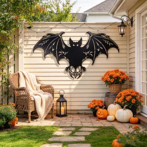 May include: A large black metal bat wall art piece with intricate cut-out wing details mounted on a light-coloured horizontal siding wall, accompanied by a wicker chair with a cream throw, a black lantern, orange mums, and several pumpkins.