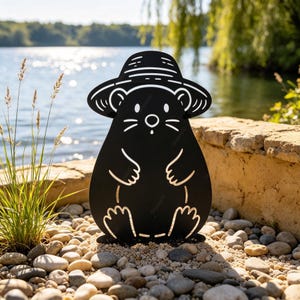 May include: A black metal silhouette of a mole wearing a wide-brimmed hat. The design features cut-out details for the eyes, nose, whiskers, mouth, and paws, creating a flat, stylised representation of the animal standing upright on a surface.