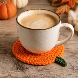 Könnte beinhalten: Ein runder, orangefarbener Häkeluntersetzer in Kürbisform mit einem kleinen grünen Häkelblatt am Rand. Eine weiße Keramiktasse, gefüllt mit einem hellbraunen Kaffeegetränk mit Schaum, steht direkt auf dem orangefarbenen Untersetzer.