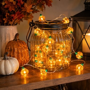 May include: A string of orange pumpkin-shaped LED lights with green stems, coiled inside a clear, ribbed glass jar with a black metal lid and handle. The illuminated pumpkin lights feature a warm glow and are connected by a thin, transparent wire.