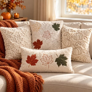 Peut inclure: Un ensemble de quatre coussins décoratifs en peluche crème ornés de broderies de feuilles d'érable et de citrouilles dans des tons orange, rouge et vert, disposés sur un canapé avec un plaid en tricot orange texturé.