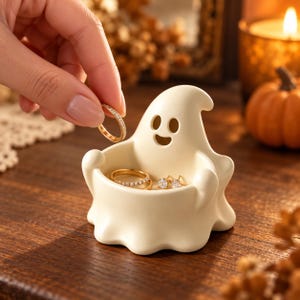 May include: A cream-colored ceramic ghost-shaped trinket dish with a hollow center for holding jewelry. The ghost figure features a smooth matte finish, a rounded head, and a smiling face with two oval eyes and a small mouth. Inside the dish sit two gold rings and a pair of small gold stud earrings.