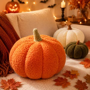 Peut inclure: Trois coussins en forme de citrouille en peluche de couleur orange, crème et vert foncé. Chaque coussin présente une surface en tissu texturé semblable à du sherpa et une petite tige distincte sur le dessus. Les citrouilles sont disposées ensemble.