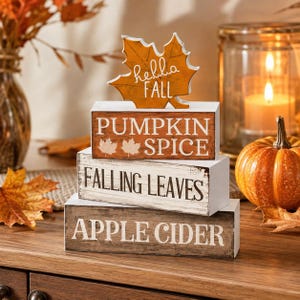 Op de afbeelding: Een stapel van vier rechthoekige houten blokken met een esdoornblad als bovenkant. Het oranje blad heeft witte tekst met hella FALL. De blokken bevatten witte tekst met PUMPKIN SPICE, FALLING LEAVES en APPLE CIDER op oranje, witte en bruine blokken.