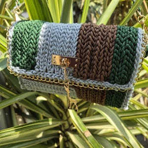 Pode incluir: Uma bolsa retangular de crochê com listras verticais em fios verde-escuro, azul-claro e marrom. A bolsa inclui um fecho de metal dourado, uma alça de corrente dourada correspondente e um pequeno pingente de girafa dourado na parte frontal.