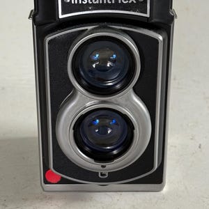 May include: A black and silver twin-lens reflex camera with the text InstantFlex printed on the front panel. The camera features two circular lenses stacked vertically, a small red dot on the lower left corner, and a separate black lens cap.