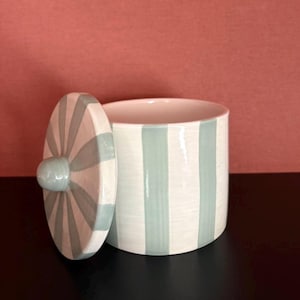 Peut inclure: Un pot cylindrique en céramique avec son couvercle assorti, orné d'un motif peint à la main de rayures verticales vert sauge clair sur fond blanc. Le couvercle présente un petit bouton de préhension arrondi au centre avec des rayures.