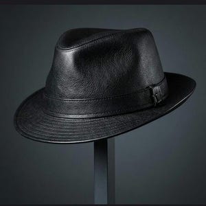 Puede incluir: Un sombrero fedora negro confeccionado en cuero texturizado. El sombrero presenta una copa pellizcada y un ala ancha. Una banda de cuero negro a juego rodea la base de la copa, asegurada con una pequeña hebilla metálica rectangular en el lateral.