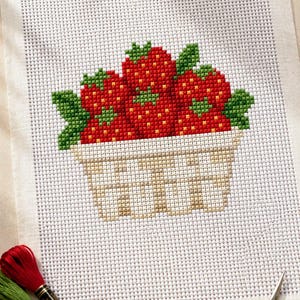 May include: A cross-stitch pattern on white Aida fabric featuring a basket filled with red strawberries and green leaves. Two skeins of red and green embroidery floss and a pair of gold-colored stork-shaped embroidery scissors are placed nearby.