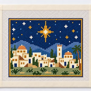 May include: A needlepoint canvas featuring a cross-stitched scene of a town with white buildings, orange domes, and a tall tower under a dark blue night sky. A large, bright yellow star shines above the landscape, surrounded by smaller white and yellow stars.