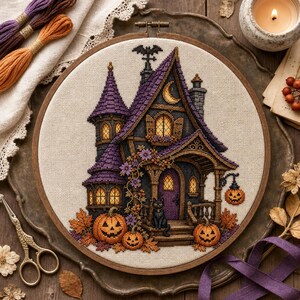 Può includere: Un ricamo a punto croce in un telaio di legno raffigurante una casa stregata viola e grigio scuro con finestre gialle luminose, un gatto nero, zucche arancioni, foglie autunnali e una luna crescente su tessuto beige chiaro.