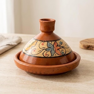 Puede incluir: Un tajine de terracota con tapa cónica que presenta una banda de color crema decorada con motivos florales pintados a mano en azul, naranja y negro. La base es un plato circular ancho y poco profundo con un acabado de arcilla marrón rojiza.