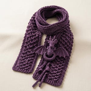 May include: A deep purple crochet scarf featuring a textured scale pattern throughout the length. Attached to the scarf is a three-dimensional crochet dragon figure with spread wings and a long tail, acting as a decorative element on the fabric.