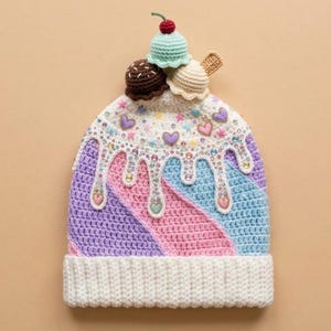 Könnte beinhalten: Eine gehäkelte Mütze mit weißem Rippenbündchen und einem Körper mit diagonalen Streifen in Lavendel, Pink und Hellblau. Die Oberseite ist mit drei kleinen gehäkelten Eiskugeln, einer Kirsche und weißem Zuckerguss mit bunten herzförmigen Perlen verziert.