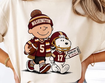 May include: A cream-colored t-shirt featuring a graphic of a cartoon character wearing a burgundy and gold beanie with the text WASHINGTON, a matching scarf, and a jersey with the number 5, holding a football next to a dog in a helmet.
