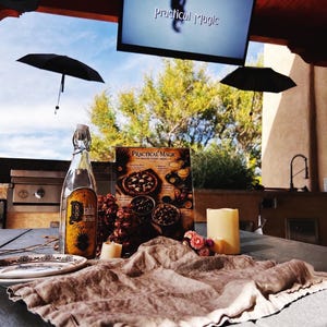 Op de afbeelding: Een tafelsetting met een fles Diablo de Flores tequila, een Practical Magic filmavond menukaart, twee crèmekleurige stompkaarsen, een klein bordje en een bruine linnen doek, onder een televisiescherm waarop de filmtitel wordt weergegeven.