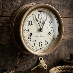 May include: A round brass wall clock with a cream-colored face, black Arabic numerals, and black hands. The dial features the text Seth Thomas and Made in U.S.A. A small brass winding key, a compass, and a coiled rope are positioned nearby.