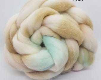 May include: A bundle of hand-dyed wool roving in shades of beige, white, and light blue. The roving is twisted together in a loose braid. The text "Theo" is written in black on a white background above the roving.