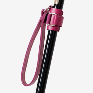 Könnte beinhalten: Ein Gehstock mit einem glänzenden, limettengrünen Derby-Griff und einem schwarzen Schaft. Eine leuchtend pinke Handschlaufe aus Leder ist mit einer passenden Manschette aus Leder und einer silbernen Metallschnalle am Schaft befestigt. Der Text DaiWalk ist sichtbar.