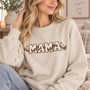Könnte beinhalten: Ein beigefarbenes Sweatshirt mit Rundhalsausschnitt und dem Wort MAMA in fetten weißen Blockbuchstaben mit dunkelbrauner Umrandung. Ein gewelltes Band mit braunem Leopardenmuster schlängelt sich durch die Buchstaben, ergänzt durch kleine rosa Sterne.