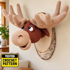 May include: A crochet moose head wall mount featuring a dark brown head, a light tan muzzle, and large tan antlers. The moose is mounted on a vertical oval-shaped slice of natural wood with bark edges. The text TESTED CROCHET PATTERN is visible.