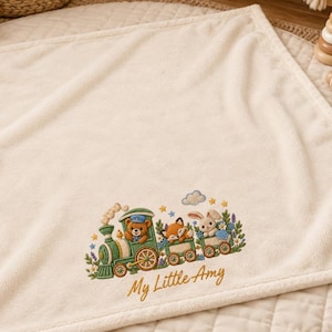 Puede incluir: Una manta de bebé cuadrada de color crema con un tren verde bordado que transporta un oso de peluche, un zorro y un conejo. El diseño incluye flores coloridas y el texto My Little Amy en letras cursivas marrones en la parte inferior.